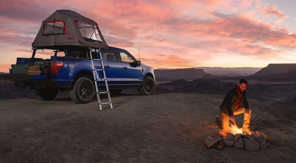 Man by campfire next to blue 2025 Ford F-150 STX FX4 with a tent on top.