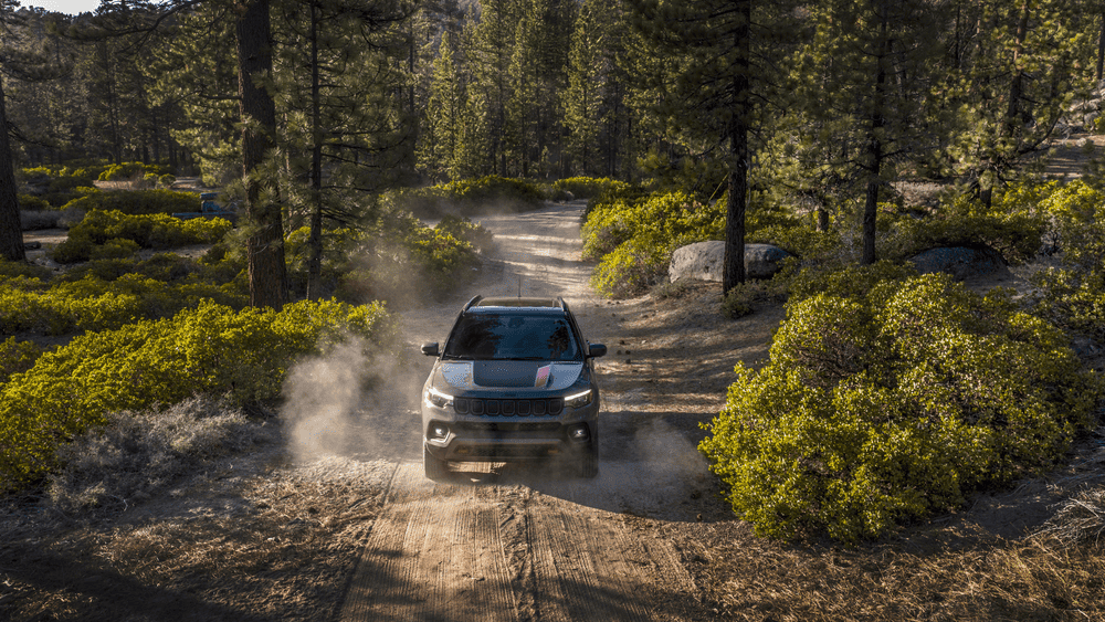 2026 Jeep Compass in Margate FL with rally stripe hood decal driving down a dusty dirt trail through a pine forest kicking up dust with LED headlights illuminated
