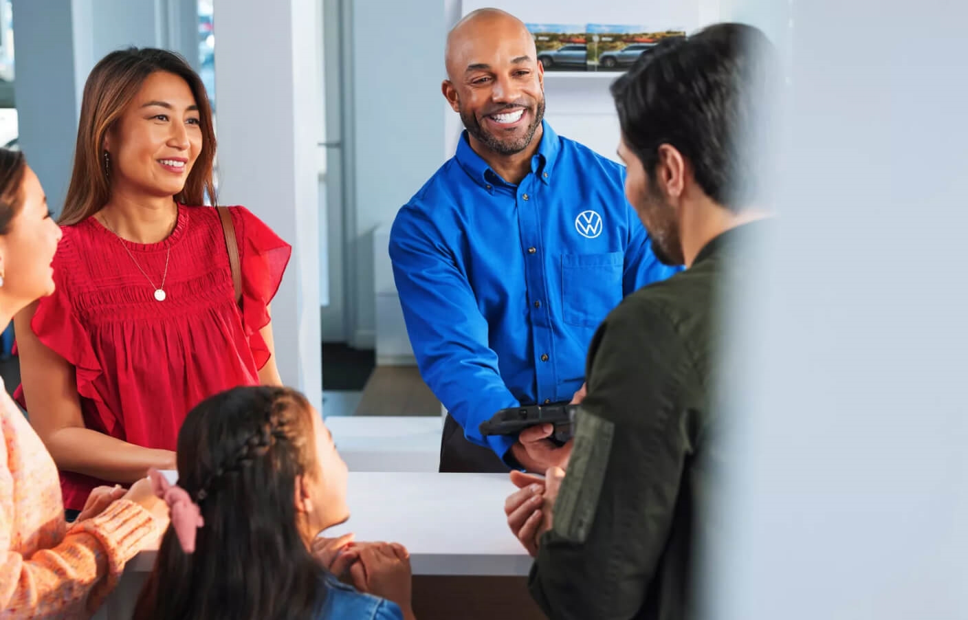 A VW Salesman holding a tablet smiles as he talks to a smiling family in a bright VW dealership.