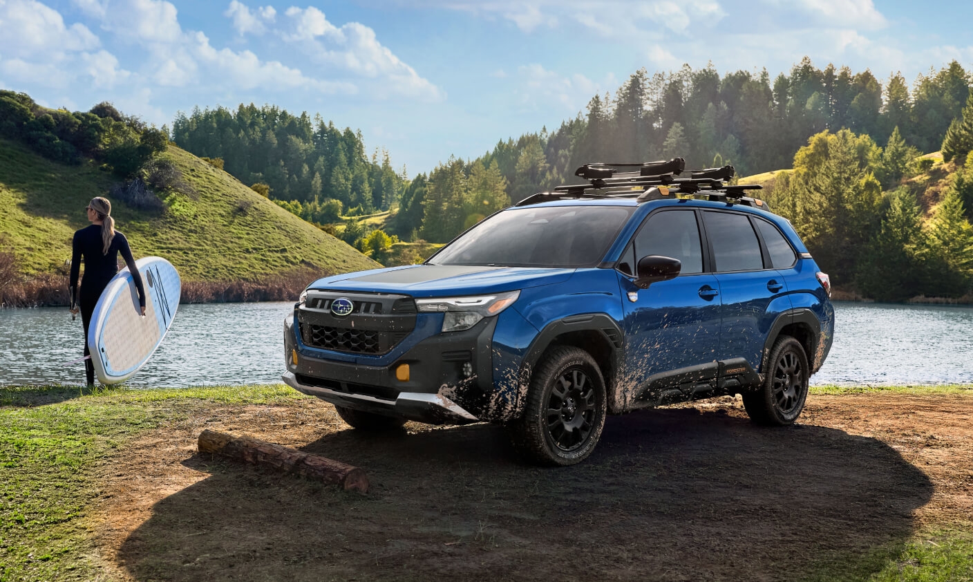 Front three-quarter view of a muddy blue 2027 Subaru Forester Wilderness by a lake with a paddleboarder.