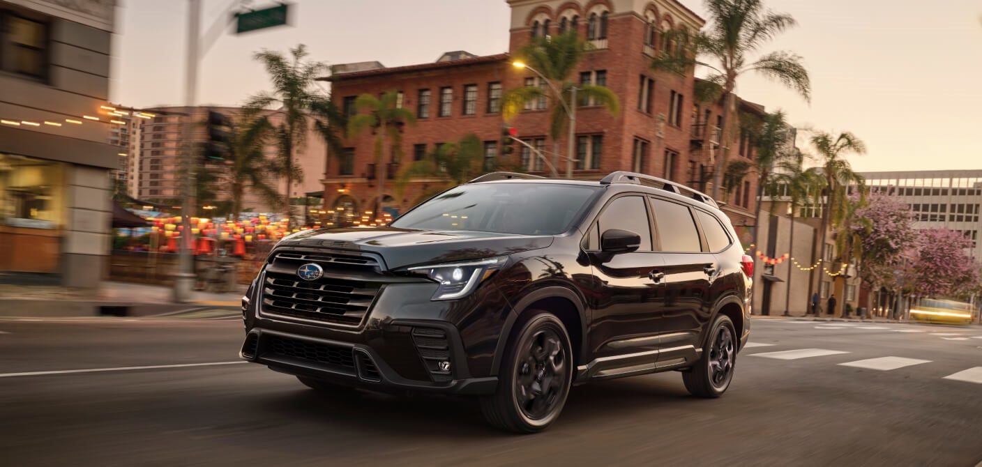Front three-quarter view of a black 2025 Subaru Ascent  driving on a city street at sunset.