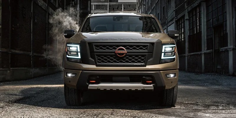 Front view of a gold Nissan Titan truck with headlights on, parked on a street with dark buildings.