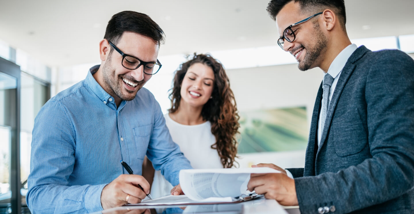 Close-up of a happy couple signing purchase paperwork with a sales representative at a car dealership.