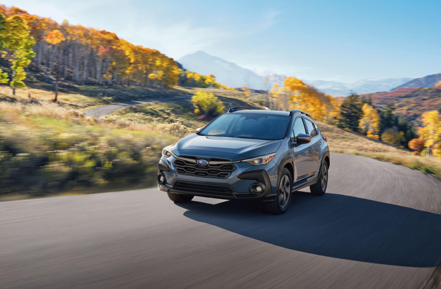 Front-angled view of a blue Subaru Crosstrek driving on a winding mountain road with autumn trees.