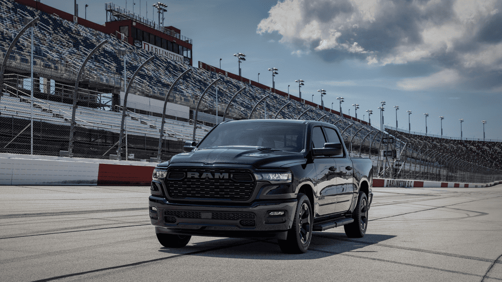 2026 Ram 1500 Express in black with blacked-out RAM grille, LED headlights, and dark alloy wheels on a NASCAR raceway — a top truck choice for Margate FL shoppers comparing it to the Ford F-150