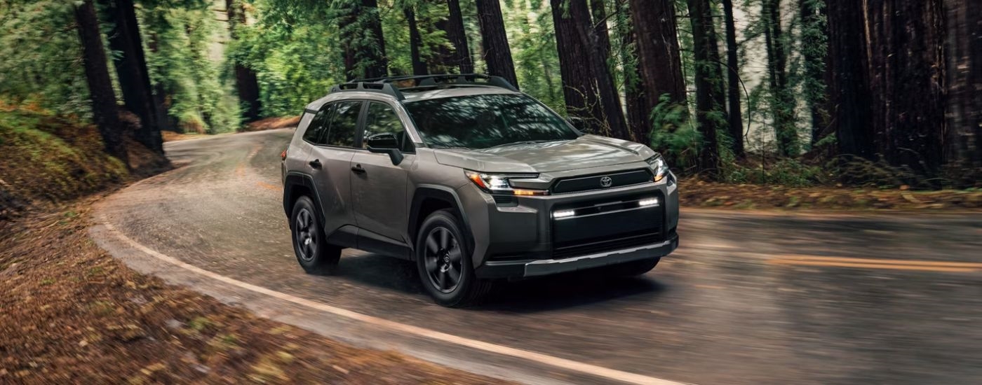 Brown 2026 Toyota RAV4 driving on a paved road through the woods.
