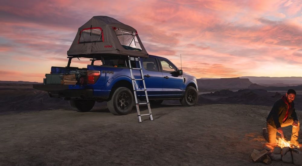 A person tending a fire next to a blue 2026 Ford F-150 STX FX4 with a tent accessory.