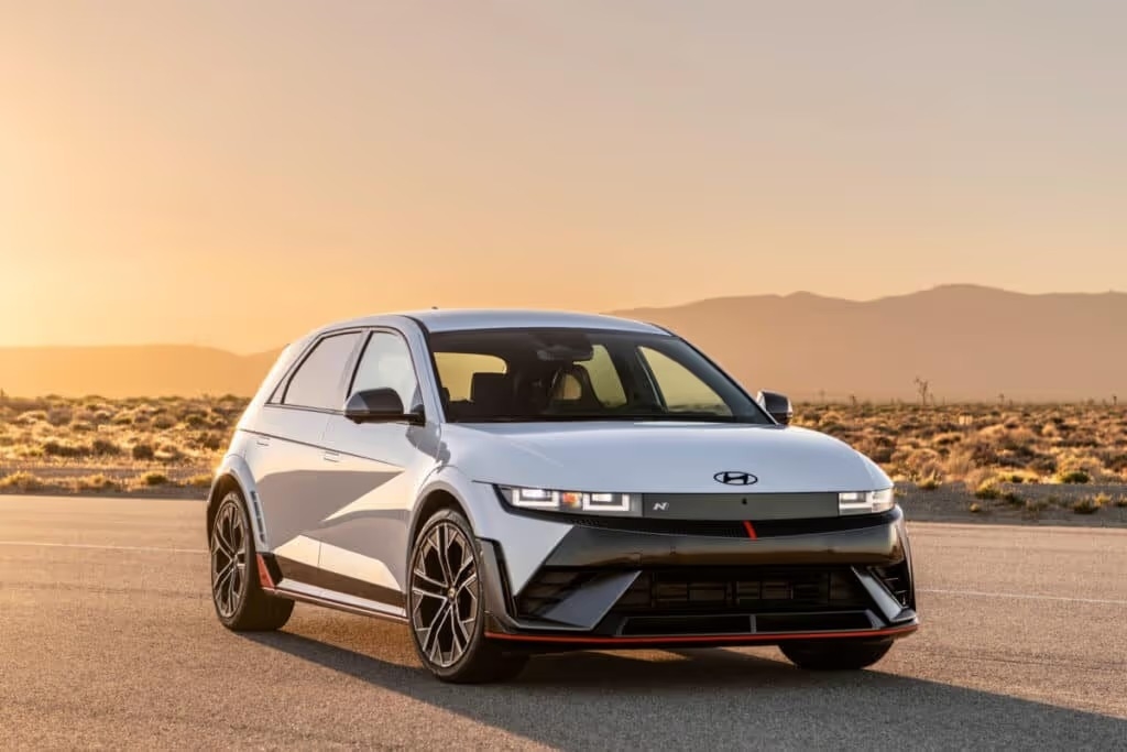 A 2025 Hyundai IONIQ 5 N SUV navigates a sunlit desert road, showcasing its sleek design against a backdrop of sandy terrain.