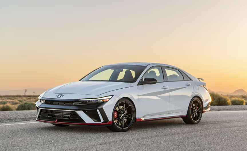 A 2025 Hyundai Elantra N drives along a scenic road during a vibrant sunset, showcasing its sleek design and performance.