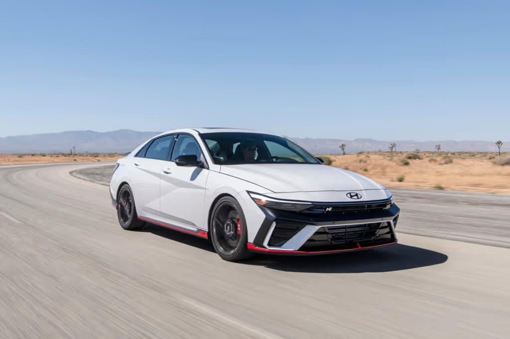 A 2025 Hyundai Elantra N drives along a sunlit desert road, showcasing its sleek design and turbocharged performance, against a vast arid landscape.