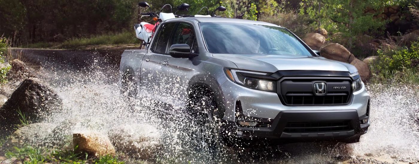 Silver 2026 Honda Ridgeline driving off-road with dirtbikes in the bed.