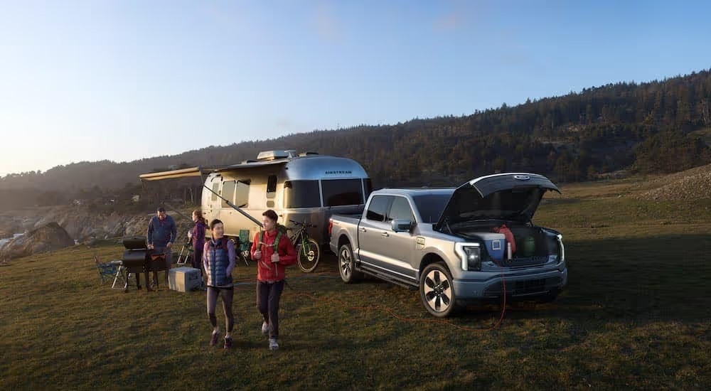 A silver 2022 Ford F-150 Lightning Platinum parked at a campsite with an Airstream trailer, and gear in the frunk