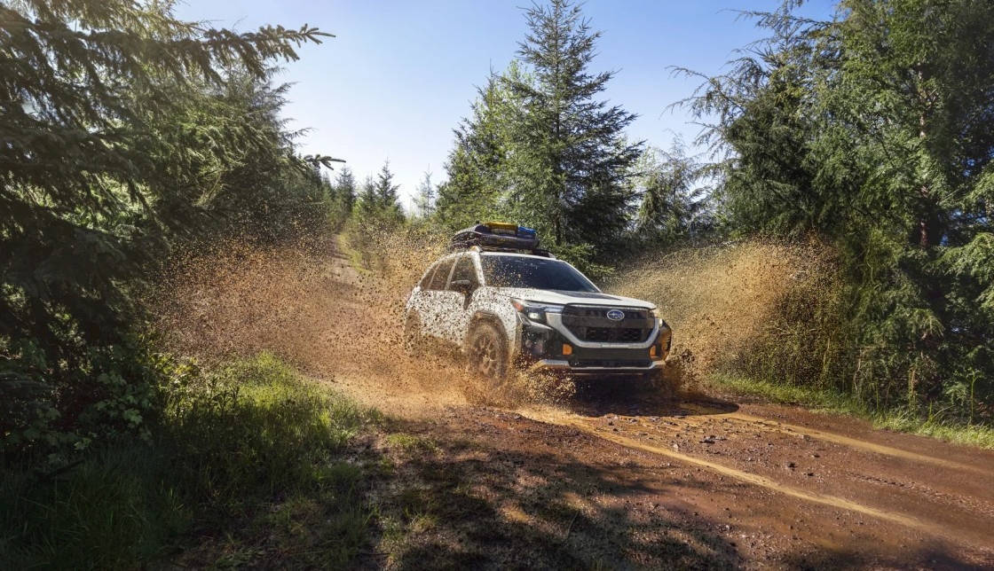 2026 Subaru Forester Wilderness charging through mud on a forest trail, cargo loaded on the roof rails, spray flying off the all-terrain tires.