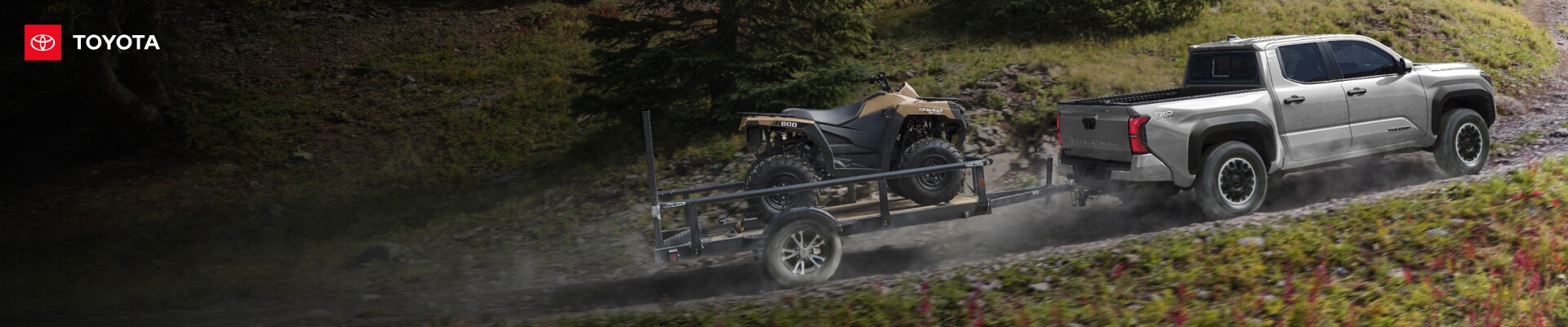 Rear-quarter view of a Celestial Silver Toyota Tacoma TRD Off-Road towing an ATV up a rugged dirt trail.
