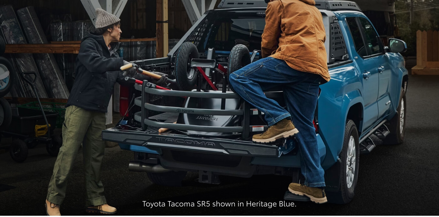Rear-quarter view of a Heritage Blue Toyota Tacoma SR5 being loaded with equipment by workers.