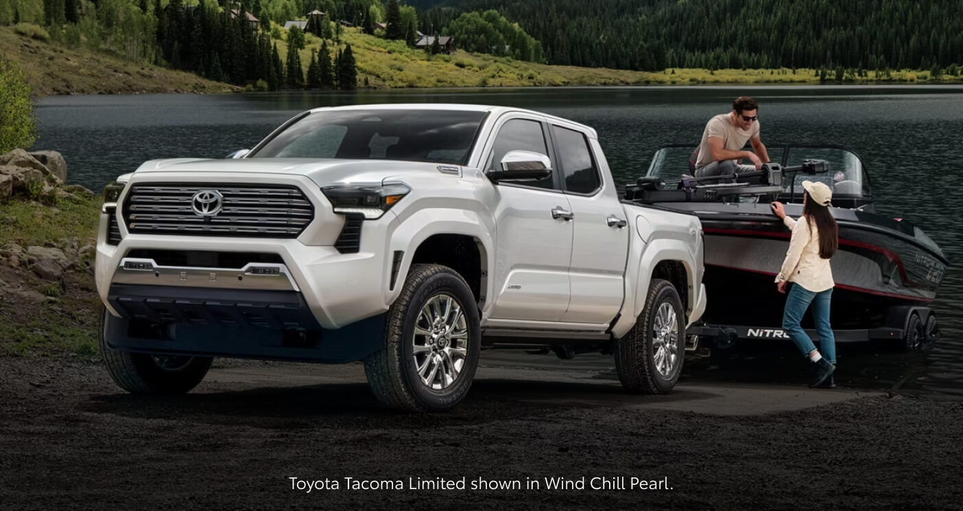 Three-quarter front view of a Wind Chill Pearl Toyota Tacoma Limited towing a boat next to a scenic lake dock.