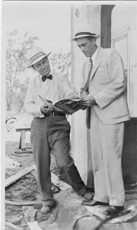 Harry Dahl in a light suit and straw hat reviewing a book with another man