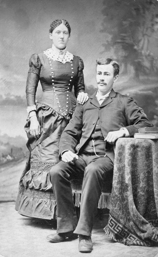 Historic studio portrait of a seated man in a suit beside a standing woman in a Victorian dress