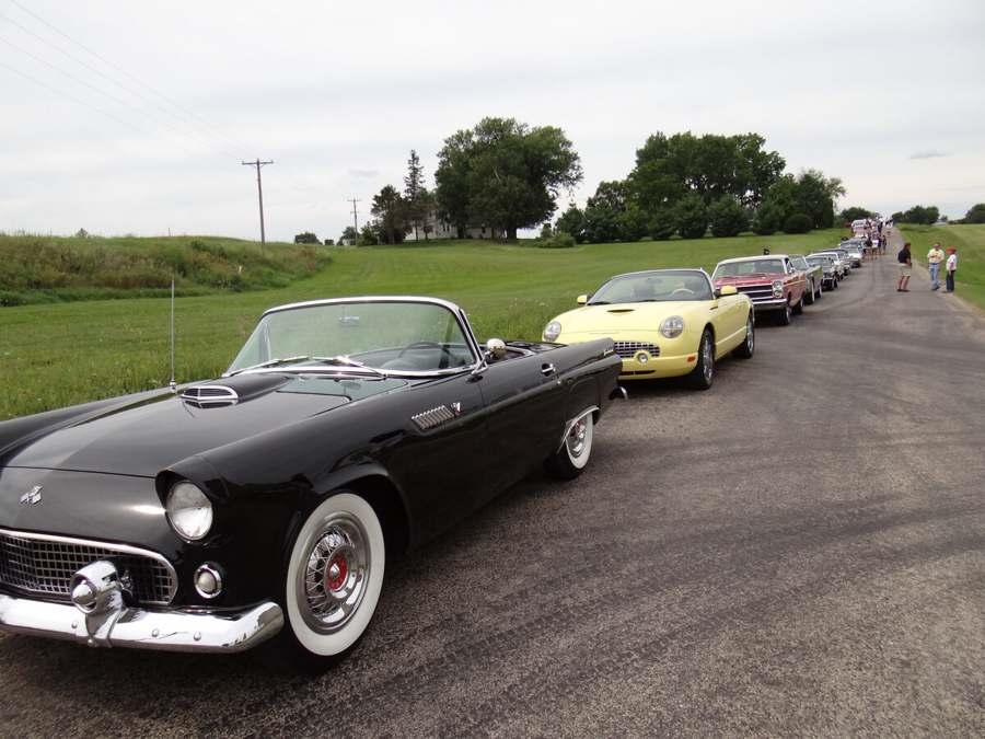 Row of classic cars on a country road with people gathered along the roadside