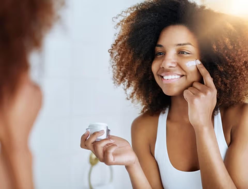A young woman applying moisturizer to her face in front of the mirror