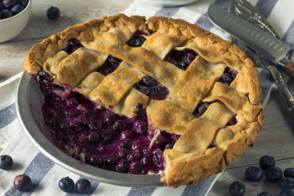 A blueberry pie with a slice missing. On a white and blue striped napkin with blueberries and serving utensils.