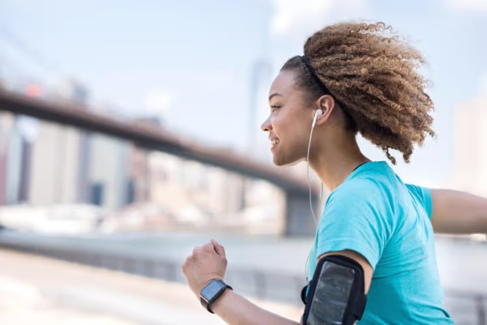 A young Black woman running outdoors in a city. She is wearing a blue shirt and an armband with her smartphone. She is wearing earbuds.