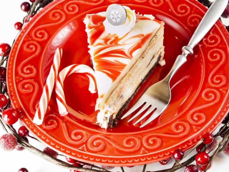 A slice of candy cane cheesecake on a festive red plate with fork and a candy cane garnish.