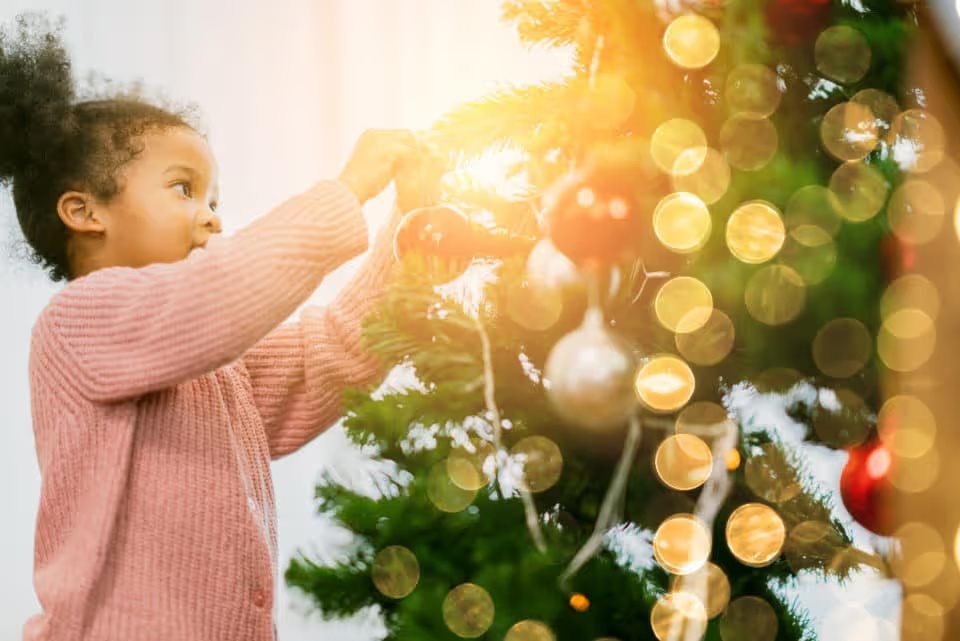 A young Black girl putting ornaments and lights on a Christmas tree.