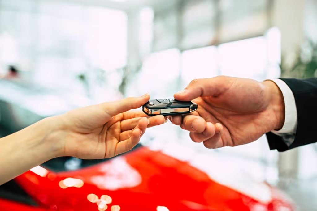 one hand giving another a car key fob in front of a blurred red car in background
