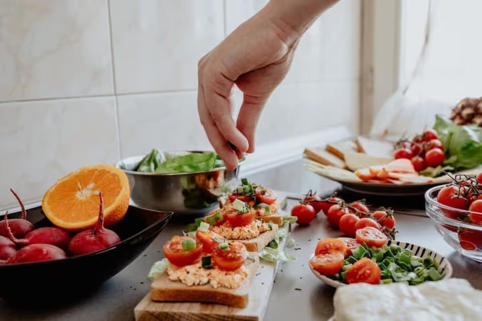 A counter full of vegetables with a vegetarian toast meal on display. A white hand is in the shot to sprinkle salt on the toast.
