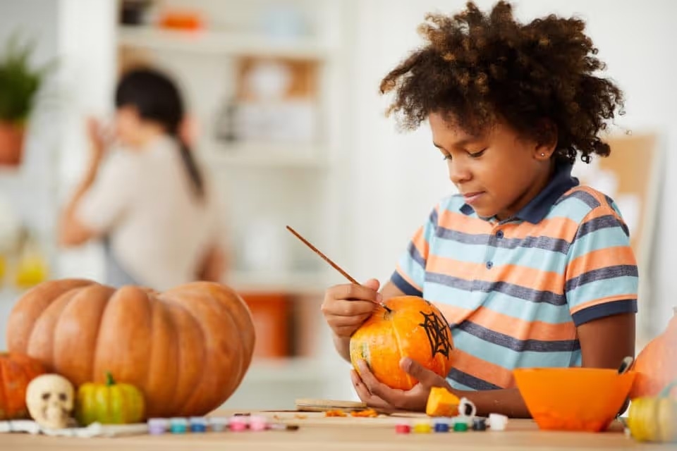 Smiling creative child leaning on counter with gouaches and making design on pumpkin