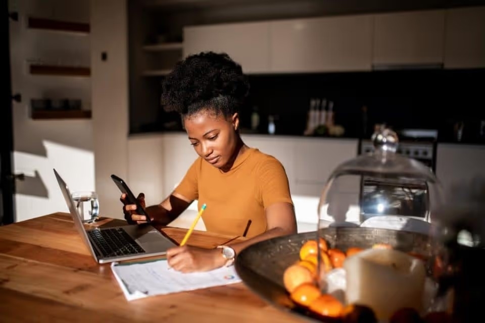 Young woman learning online with laptop and phone, she is taking notes in a notebook