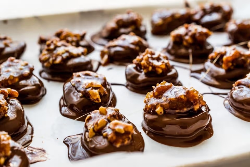 A tray of freshly made chocolate turtle candies. The chocolate is still wet and shiny and there are walnuts on top of each candy mound. There are 16 candy mounds shown.