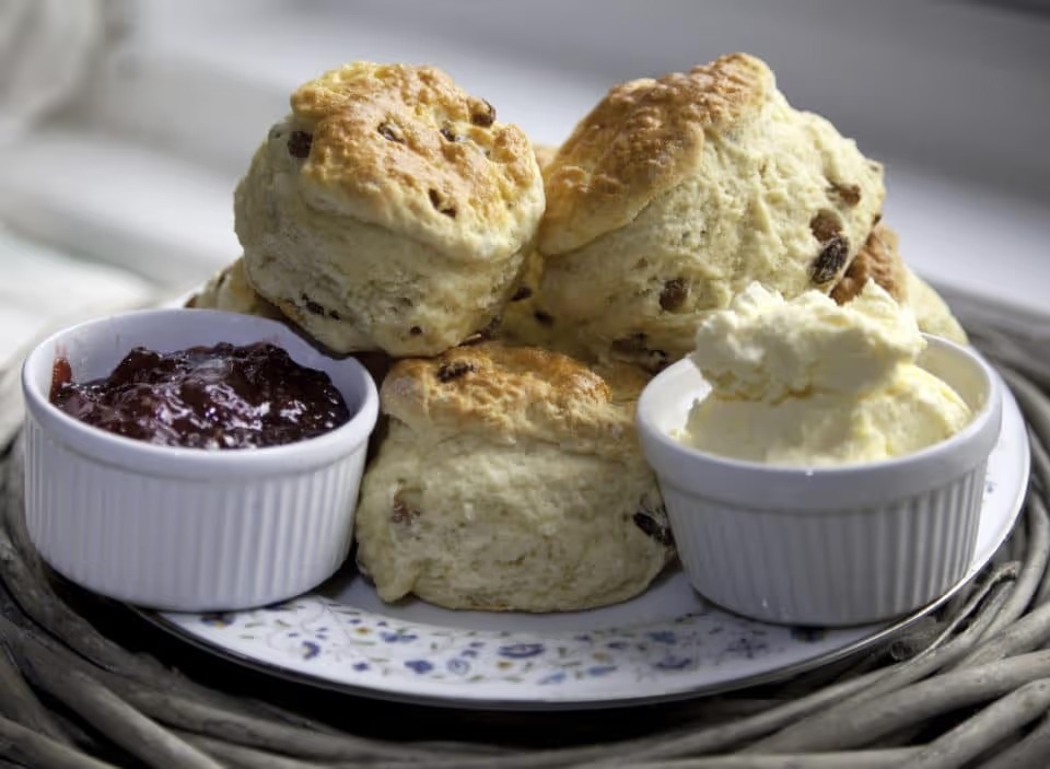 Several scone stacked on a porcelain plate with ramekins filled with raspberry jam and clotted cream.