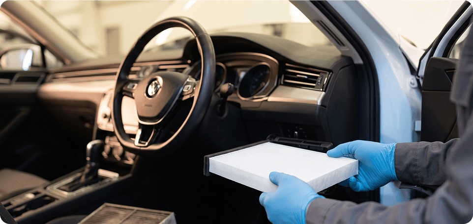 Gloved technician holding a new cabin air filter inside a Volkswagen, with the used filter below the dashboard