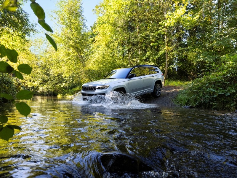 A gray 2026 Jeep Grand Cherokee drives int oa creek in a wooded landscape.