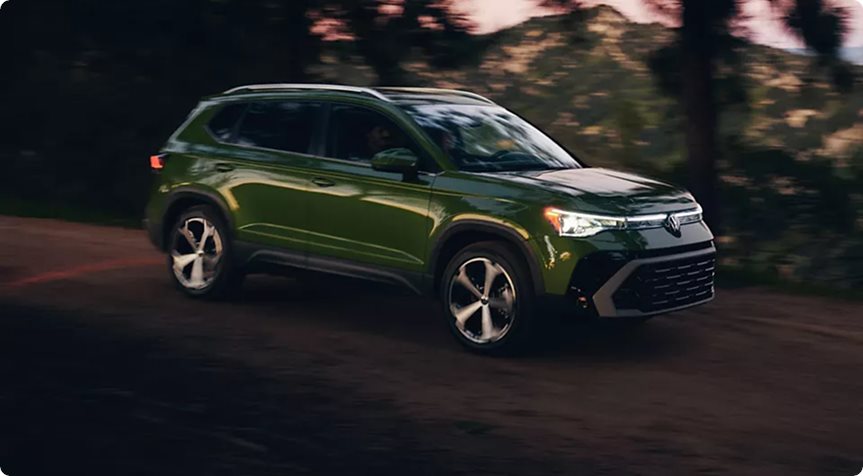 Green 2027 Volkswagen Taos driving through the woods at dusk with its LED headlights on