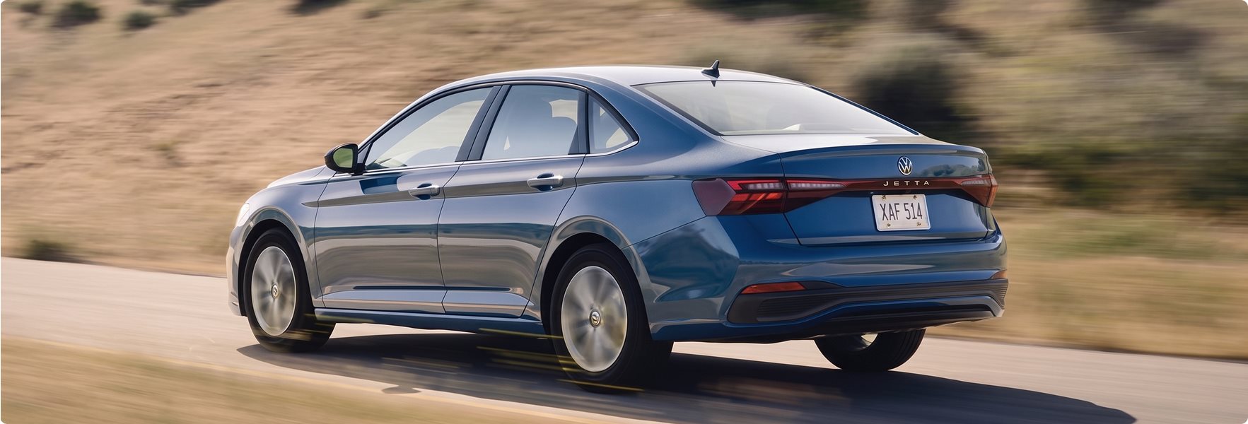 Blue 2027 Volkswagen Jetta driving past dry hillsides, seen from the rear