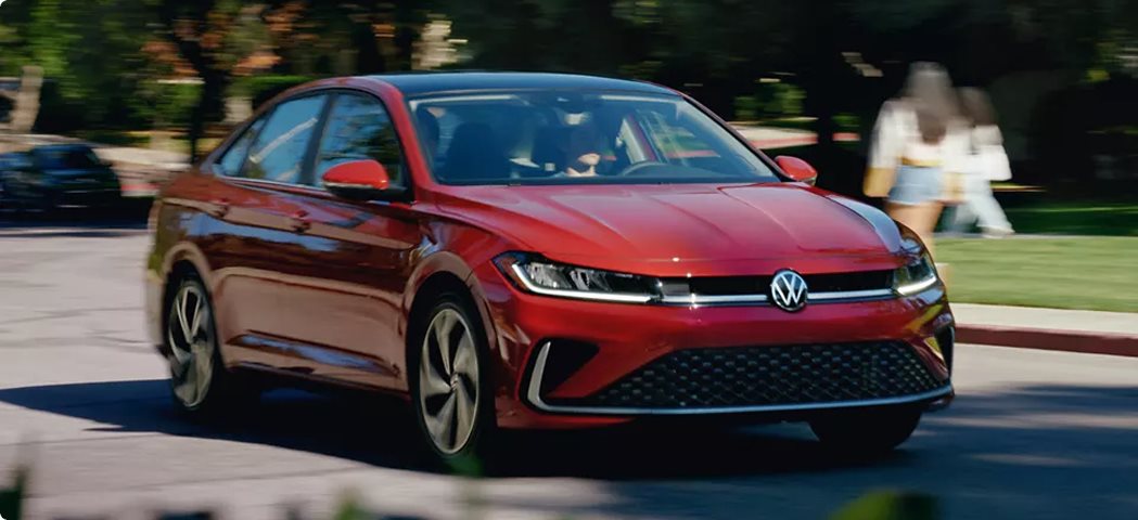 Red 2027 Volkswagen Jetta with its LED headlights on, driving down a tree-lined street