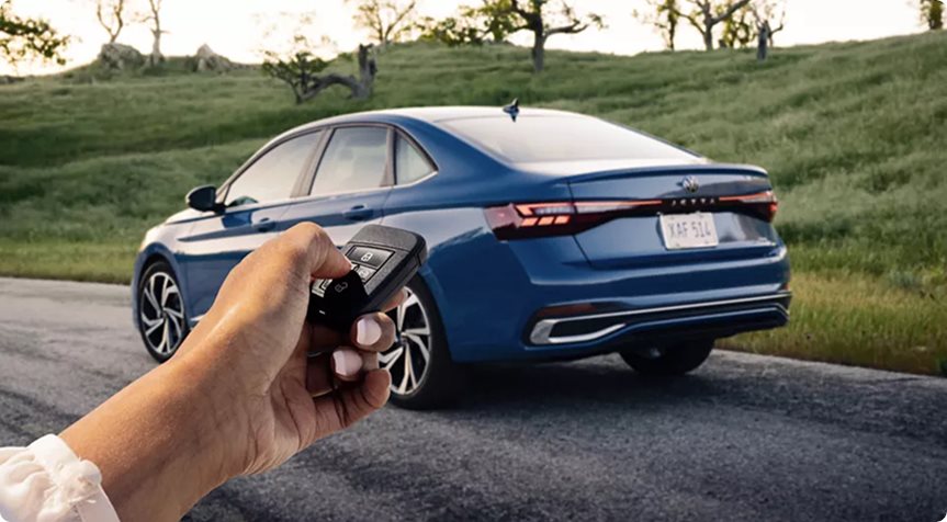 Hand holding a Volkswagen key fob behind a blue Jetta parked beside a grassy hill