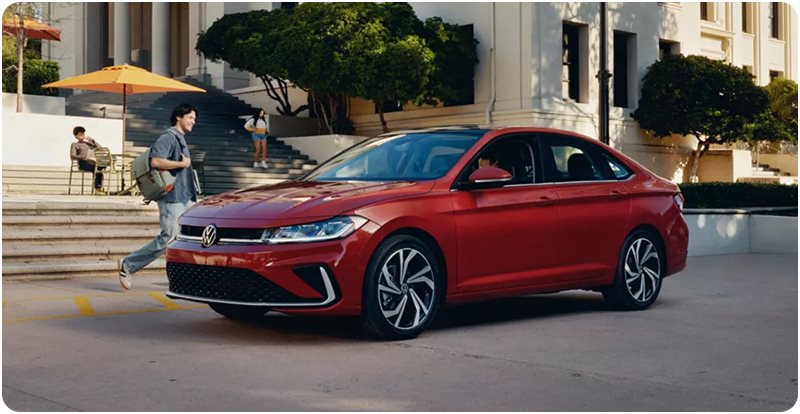 Red Volkswagen Jetta parked outside a campus building as students walk by