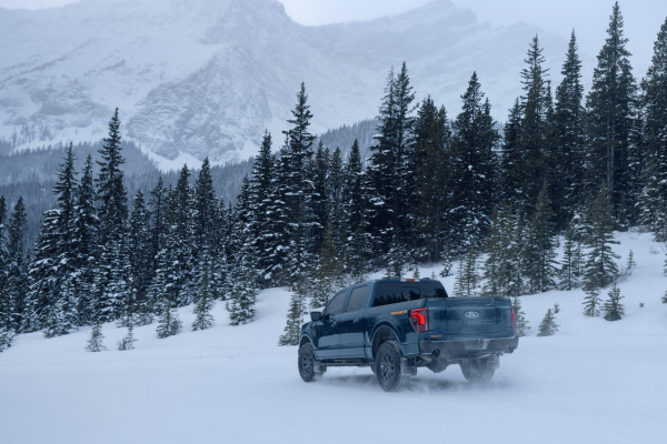 Ford F-150 driving through deep snow in a mountainous winter landscape surrounded by snow-covered evergreen trees.