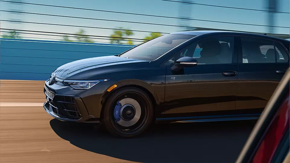 Side view of a dark 2026 Volkswagen Golf R driving past a stadium