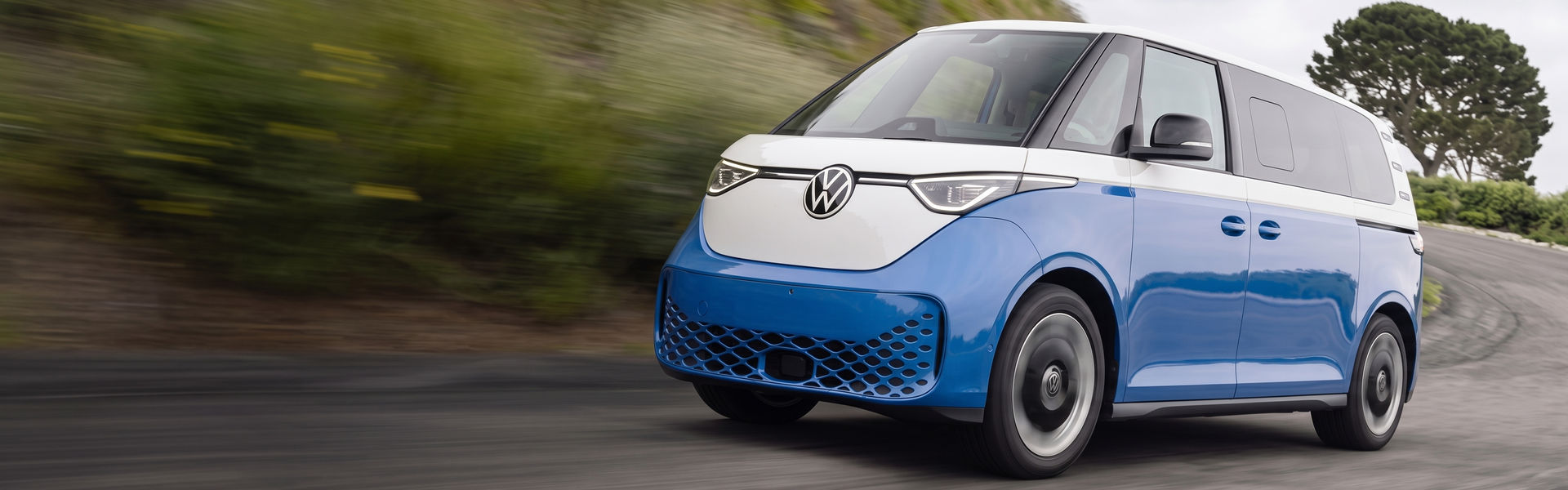 Blue and white 2025 Volkswagen ID. Buzz driving along a winding coastal road