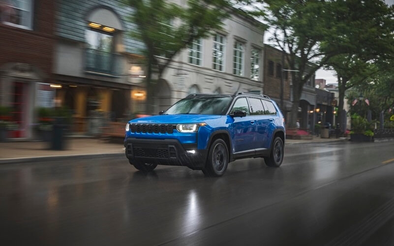 A blue 2026 Jeep Cherokee drives down a main street.