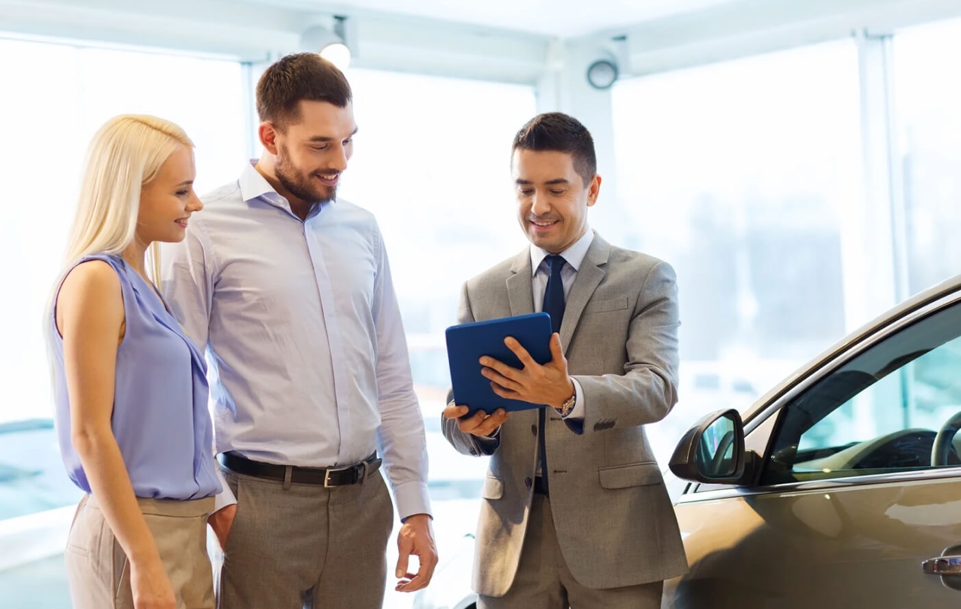 A sales representative shows a pair of smiling customers a tablet in a bright dealership.