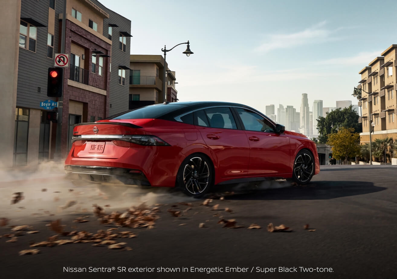 Rear three-quarter view of a 2027 Nissan Sentra SR in Energetic Ember and Super Black kicking up autumn leaves on an urban street.