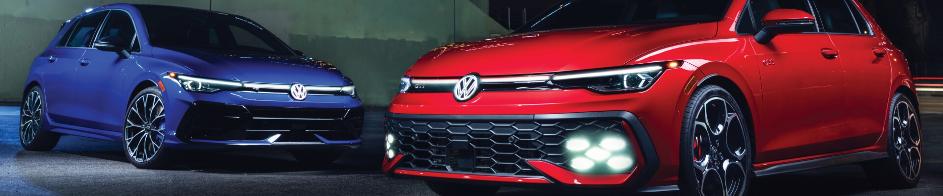 Front three-quarter view of a blue Volkswagen Golf R and red Golf GTI on dark pavement.