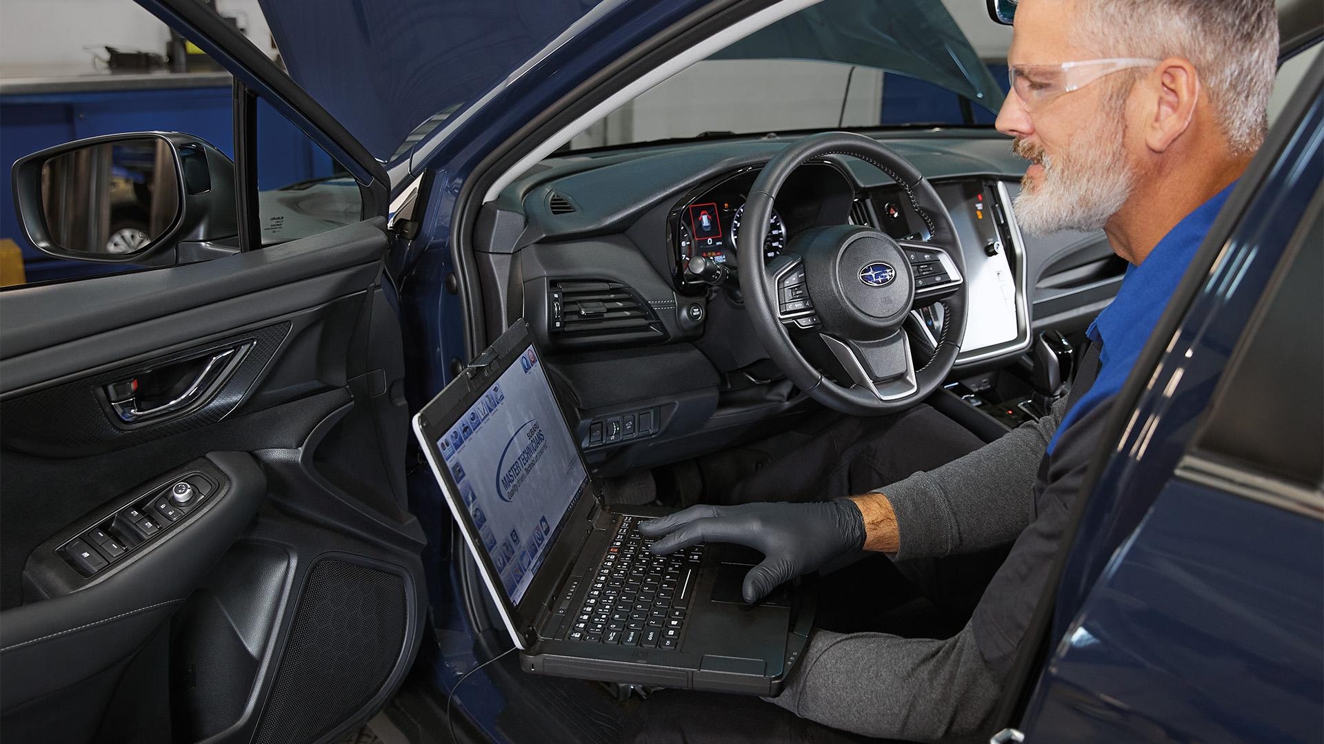 A Subaru Master Technician working at a laptop in the driver's seat of a blue Subaru in the service bay
