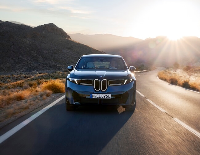 A front view of a 2027 BMW iX3 driving on a mountain highway at sunset.
